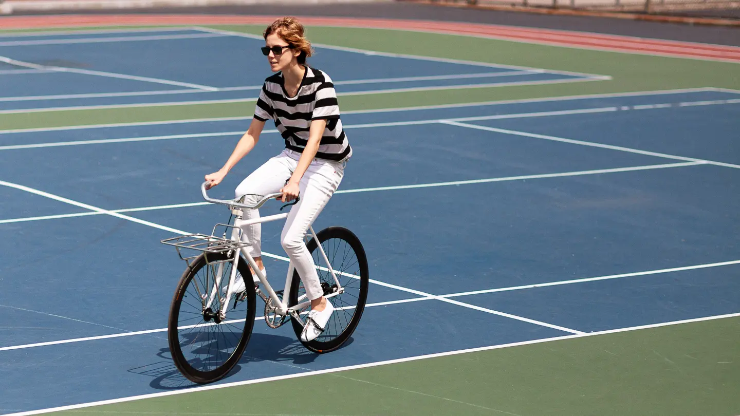 Art Historian, Claudia Eve Beauchesne rides a bike on a tennis court, wearing sunglasses