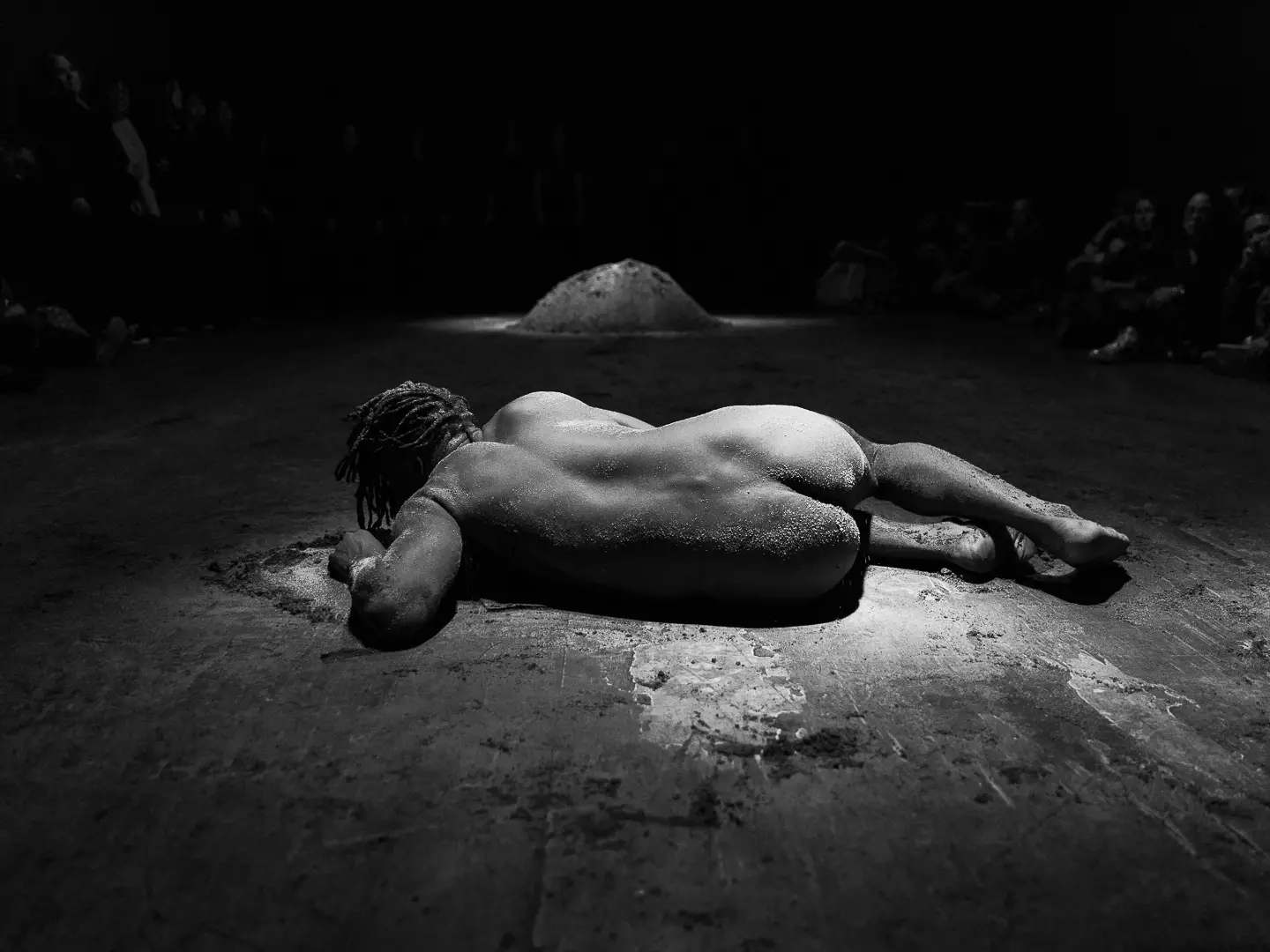 Carlos Martiel lies face down, partially covered in a dirt, on a dimly lit surface.